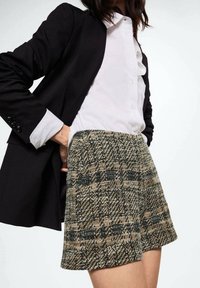 Shorts a vita alta con motivo a quadri, nelle tonalità di beige e nero, realizzati in tessuto testurizzato; abbinati a una camicia bianca con bottoni e a un blazer nero.
