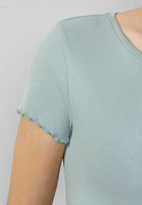 Nahaufnahme eines gerippten hellblauen Kurzarmhemds mit einem Blütenblattabschluss am Ärmel, getragen von einer Person.
