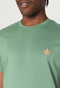 Grön bomullströja med en liten broderad sköldpaddedesign i beige färg, med klassisk rund hals och mjuk textur.