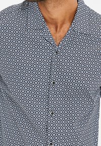 Chemise à manches courtes bleu marine avec un motif géométrique en diamants blancs et bleu clair, dotée d'un col et de boutons noirs.