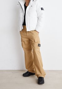 Giacca a vento bianca con cappuccio, dettagli neri e logo. Pantaloni cargo color cammello con tasche laterali e toppa con logo. Scarpe da ginnastica nere.
