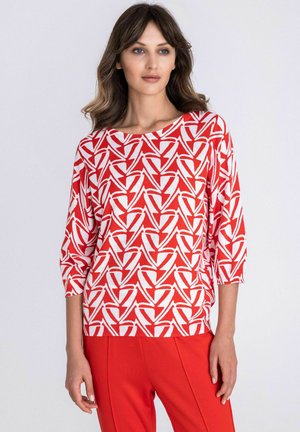 Vrouw met halflang golvend haar draagt een top met rood-wit geometrisch patroon, driekwart mouwen en effen rode broek.