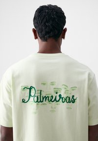 Camisa de algodón verde claro con mangas cortas, que presenta un gráfico de rostros y la palabra "Palmeiras" en cursiva verde más oscuro en la parte posterior.
