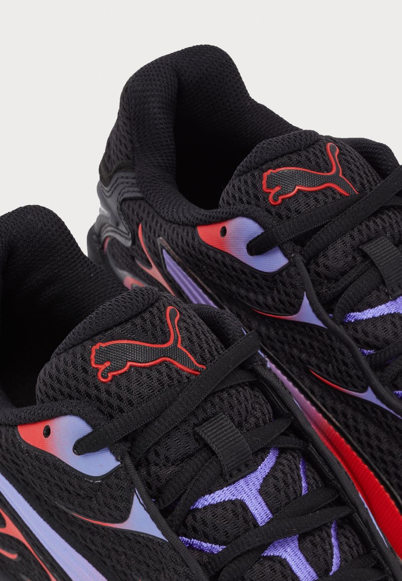 Zapatillas de running de malla negra con acentos en rojo y púrpura, que cuentan con superficies texturizadas, lazadas finas y un logo prominente de Puma en la lengüeta.