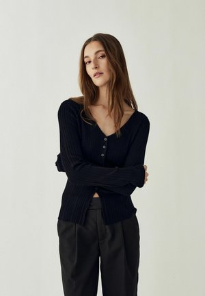 Mujer joven con cabello largo y castaño, vestida con un cárdigan negro acanalado con botones y pantalones negros, de pie con los brazos cruzados frente a un fondo liso.