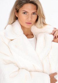 Donna con capelli biondi che indossa un cappotto di peluche bianco, tiene il collare con una mano e guarda direttamente verso la macchina fotografica.