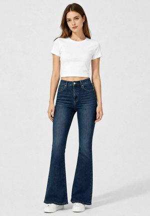 Young woman standing wearing a white cropped t-shirt, dark blue high-waisted flared jeans, and white sneakers against plain background.