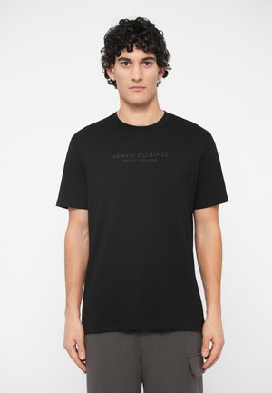 Man in een zwart Armani Exchange T-shirt met de tekst Milano/New York, staand tegen een effen witte achtergrond, recht vooruit kijkend.