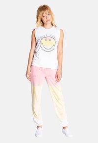 Witte mouwloze top met een glimlachende graphic, gecombineerd met roze-naar-geel verloop joggingsbroeken met kleine glimlachprints, en witte schoenen.