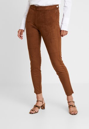Femme portant un pantalon en daim marron ajusté avec une fermeture éclair latérale, un chemisier blanc à manches longues et des sandales à talons imprimé léopard, debout sur un sol blanc.
