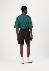 Πράσινο κοντομάνικο πουκάμισο με μαύρες λεπτομέρειες σε συνδυασμό με μαύρες βερμούδες με ελαστική μέση και πλαϊνή τσέπη, φορεμένο με λευκά αθλητικά παπούτσια.