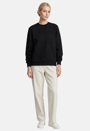 Mujer con cabello rubio con una sudadera negra, pantalones beige claros y zapatillas blancas, de pie contra un fondo blanco liso.