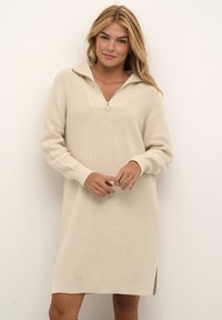 Stickad beige klänning med halvdragkedja, långa ärmar, sidokilar samt ribbade mudd och nederkant. Texturerad tyg med avslappnad passform.