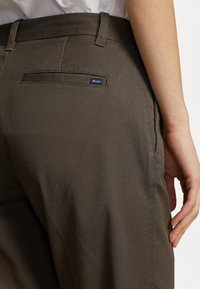 Närbild av en persons midja iförd mörkbruna Polo-byxor med bältesöglor och en liten bakficketikett, hand vilande vid sidan.