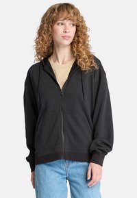 Schwarze Reißverschluss-Hoodie mit Kordeln, Fronttaschen und gerippten Bündchen, getragen über einem hellen, beige Shirt, kombiniert mit hellblauen Jeans.