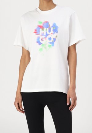 Mujer con camiseta blanca con estampado floral colorido y borroso y texto "HUGO", combinada con pantalones negros sobre un fondo liso.