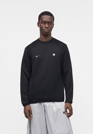 Hombre que lleva una sudadera negra de manga larga de Nike y pantalones blancos holgados, de pie contra un fondo liso y claro.