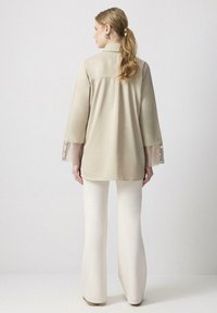 Chemise beige à manches longues avec un léger éclat, présentant un empiècement au dos et des accents en tissu transparent sur les manches. Associée à un pantalon blanc évasé.
