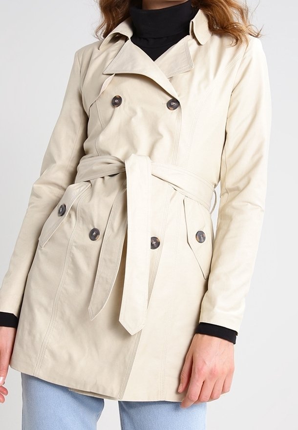 Trench-coat beige à double boutonnage avec ceinture à la taille, porté sur un col roulé noir et un jean bleu clair, visible des épaules aux hanches.