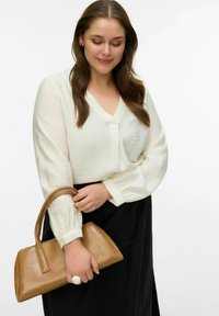 Blouse en ivoire texturée à manches longues et col en V, associée à une jupe noire. Tenant un petit sac à main en cuir marron.