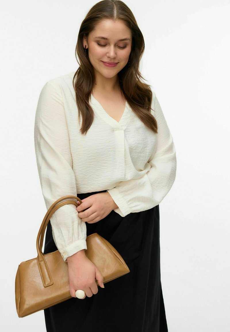 Blouse en ivoire texturée à manches longues et col en V, associée à une jupe noire. Tenant un petit sac à main en cuir marron.