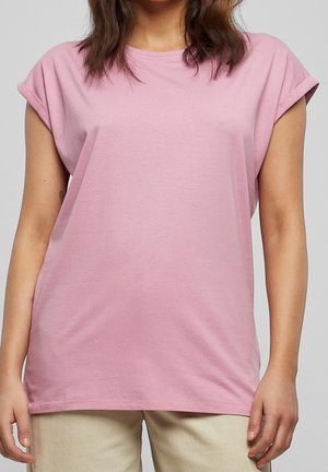 Vrouw draagt een lichtroze shirt met korte mouwen met opgerolde mouwen, beige broek, staand tegen een effen grijze achtergrond.