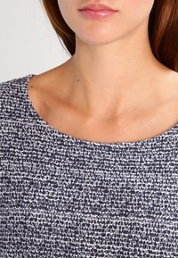 Marineblau und weißer strukturierter Stoffoberteil mit weitem, abgerundetem Ausschnitt. Die Webart zeigt ein gemischtes Muster, das der Gestaltung Tiefe verleiht.