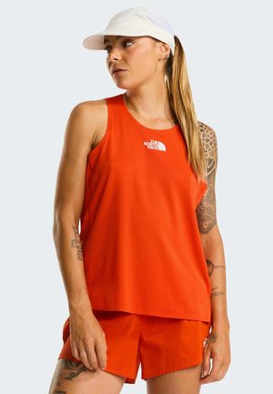 Mujer con gorra blanca, top sin mangas naranja y pantalones cortos a juego, con tatuajes visibles en brazos y pierna, mirando de lado.