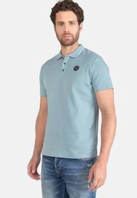 Lichtblauw polo shirt met korte mouwen en een kraag, met een drie-knops sluiting en een logo op de linkerkant van de borst. Gedragen met blauwe spijkerbroek.