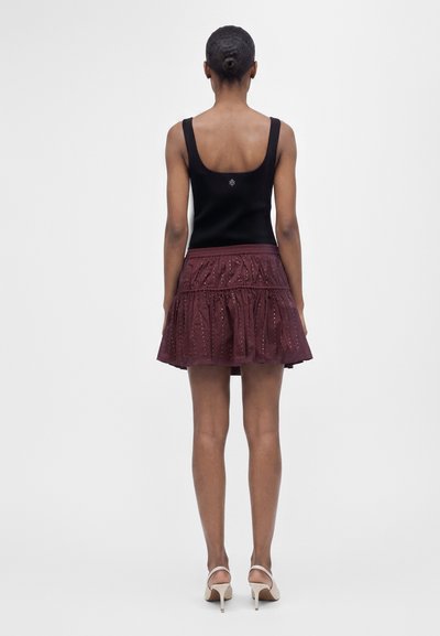 Femme avec cheveux en chignon, portant un haut noir sans manches, une jupe à volants violette ornée de sequins et des sandales blanches à talons, debout face à l'arrière.