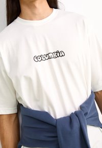 Camiseta blanca con cuello redondo y mangas cortas, que presenta un logo en letras de burbuja negras que dice "COLUMBIA". Un suéter azul atado a la cintura.