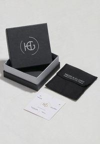 Black box with silver logo, grey interior, containing a black pouch and two tags on a light fabric surface. Pouch displays white text.