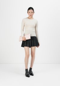 Kvinne iført beige teksturert langermet topp, svart plissert skjørt, grå sokker, svarte sko, og holder en lys rosa clutchtaske mot en hvit bakgrunn.