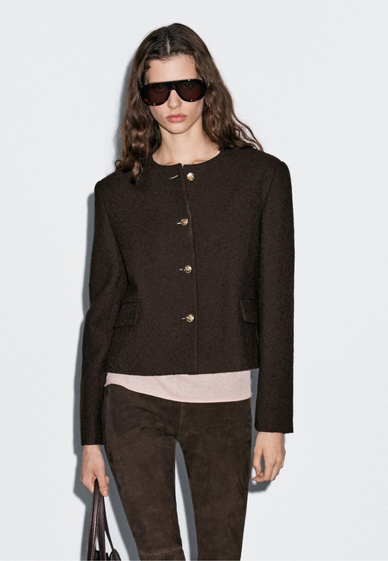 Vrouw in een oversized donkerbruine blazer met gouden knopen, bijpassende broek, lichtroze top, grote donkere zonnebril, en houdt een donkere tas vast.