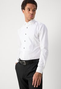 Fehér ing, hegyes gallérral, hosszú ujjal és fekete gombokkal díszített elülső részen; fekete öltönynadrággal és övvel párosítva.