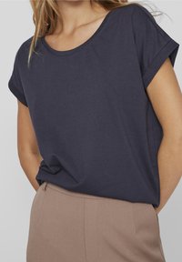 Žena v tmavě modrém tričku s krátkým rukávem, zastrčeném do kalhot s vysokým pasem v světle hnědé barvě, stojí na pozadí bez vzoru.