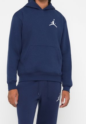 Person trägt dunkelblauen Hoodie und passende Jogginghose mit weißem Jumpman-Logo auf Brust und oberem Oberschenkel.
