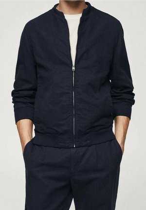 Homme portant une veste zippée bleu marine sur une chemise blanche, avec un pantalon assorti bleu marine et les mains dans les poches.