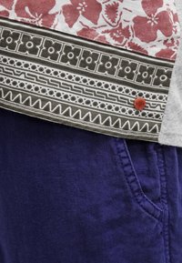Korte mouwen shirt met rode en grijze bloemmotieven; geprinte zoom en een bruine accentknop; gecombineerd met diep paarse broek.