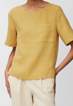 Osoba ubrana w luźną, musztardowożółtą koszulę z krótkim rękawem i kieszenią na piersi oraz jasnobeżowe spodnie, stojąca z rękami w kieszeniach.