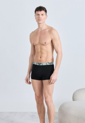 Noor mees seisab plaanilisel heledal taustal mustade bokserpükstega, millel on hall kaubamärgiga vöö.