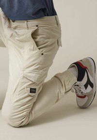Pantalons cargo beiges avec plusieurs poches, boutons et une finition texturée. Portés avec des baskets blanches et grises avec des accents rouges et verts.