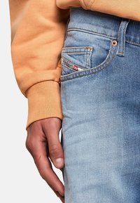 Vaqueros de denim azul claro con una textura desgastada. Manga de sudadera naranja visible, mano reposando en los vaqueros. El bolsillo presenta una etiqueta roja y blanca.