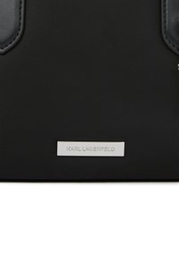 Μαύρη υφασμάτινη τσάντα χειρος με ασημένια ορθογώνια πινακίδα που φέρει χαραγμένο το "KARL LAGERFELD" κεντραρισμένο κοντά στο κάτω μέρος.