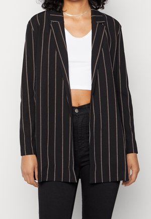 Vrouw draagt een zwarte blazer met witte verticale strepen over een wit croptop en zwarte broek, staand tegen een effen achtergrond.