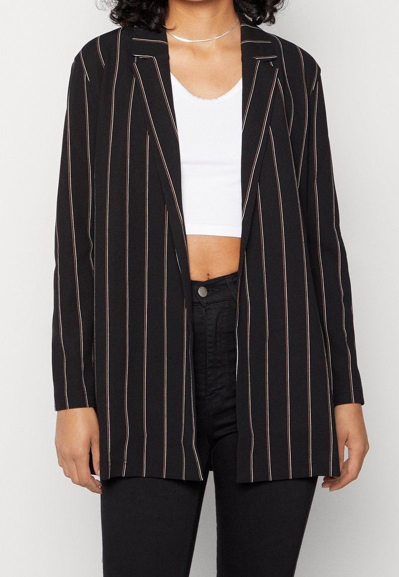 Femme portant un blazer noir à rayures verticales blanches sur un crop top blanc et un pantalon noir, debout devant un fond uni.