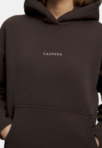 Brun hoodie med frampocket, ribbade ärmslut och dragsko i huvan. "CASPARA"-logotyp tryckt i vitt på bröstet. Mjukt material.