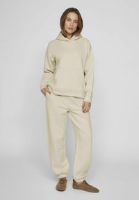 Pantalones de chándal beige y sudadera con capucha a juego. Textura suave, corte relajado, bolsillo frontal en la sudadera, tobillos ajustados en los pantalones.