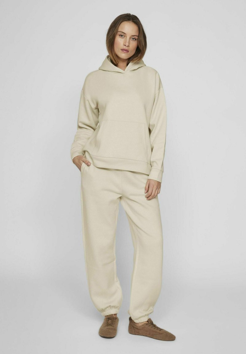Pantalones de chándal beige y sudadera con capucha a juego. Textura suave, corte relajado, bolsillo frontal en la sudadera, tobillos ajustados en los pantalones.