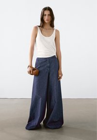 Loose-fitting dark blue denim pants with a high waist, styled with a white tank top and a small woven brown handbag.